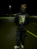 Person wearing black hoodie with a graphic of a ronin resting against a tree and a red rising sun