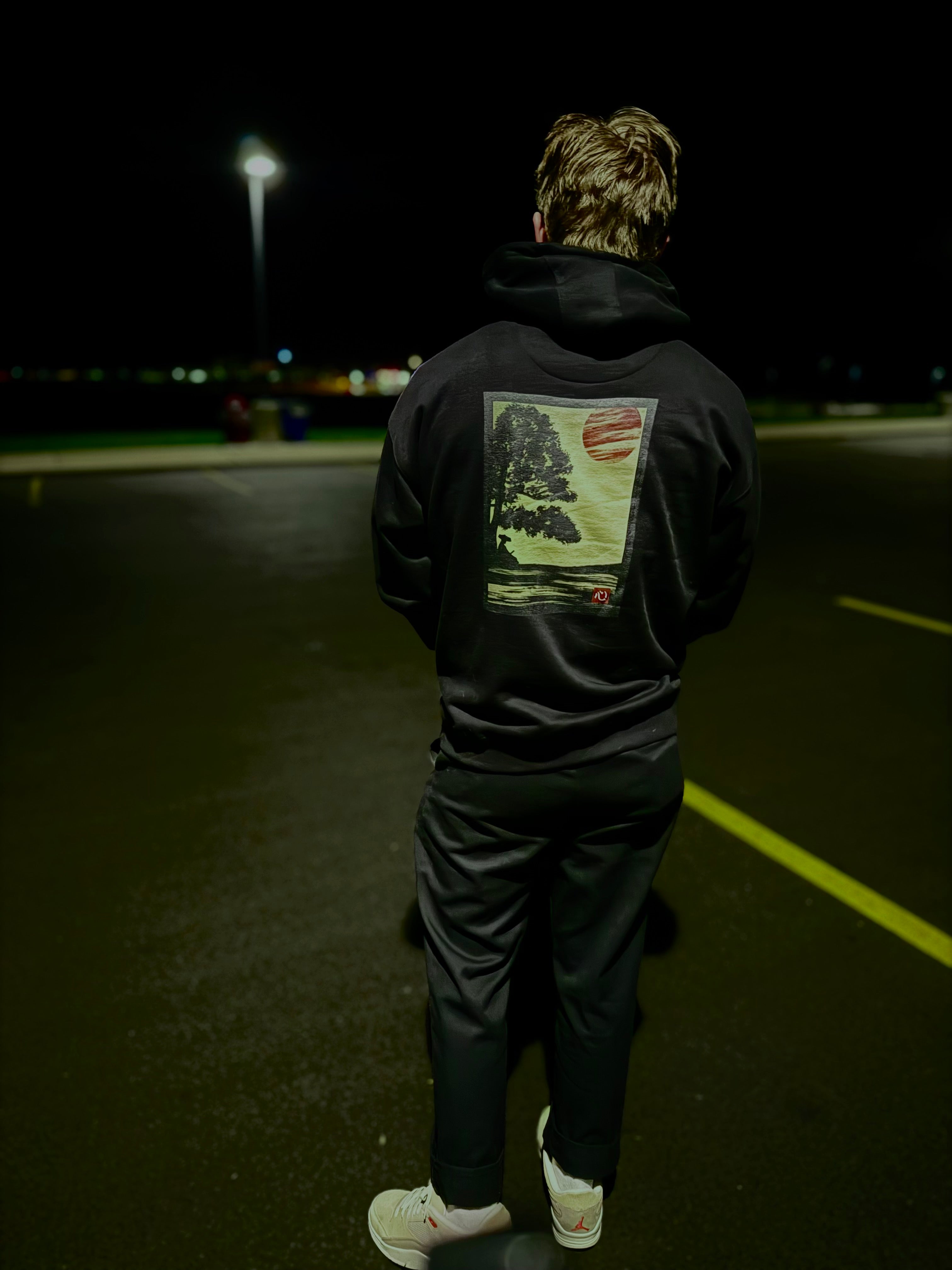 Person wearing black hoodie with a graphic of a ronin resting against a tree and a red rising sun