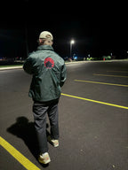 Person in at night in an empty parking lot wearing a lightweight green windbreaker with Japanese red sun and samurai helmet embroidery