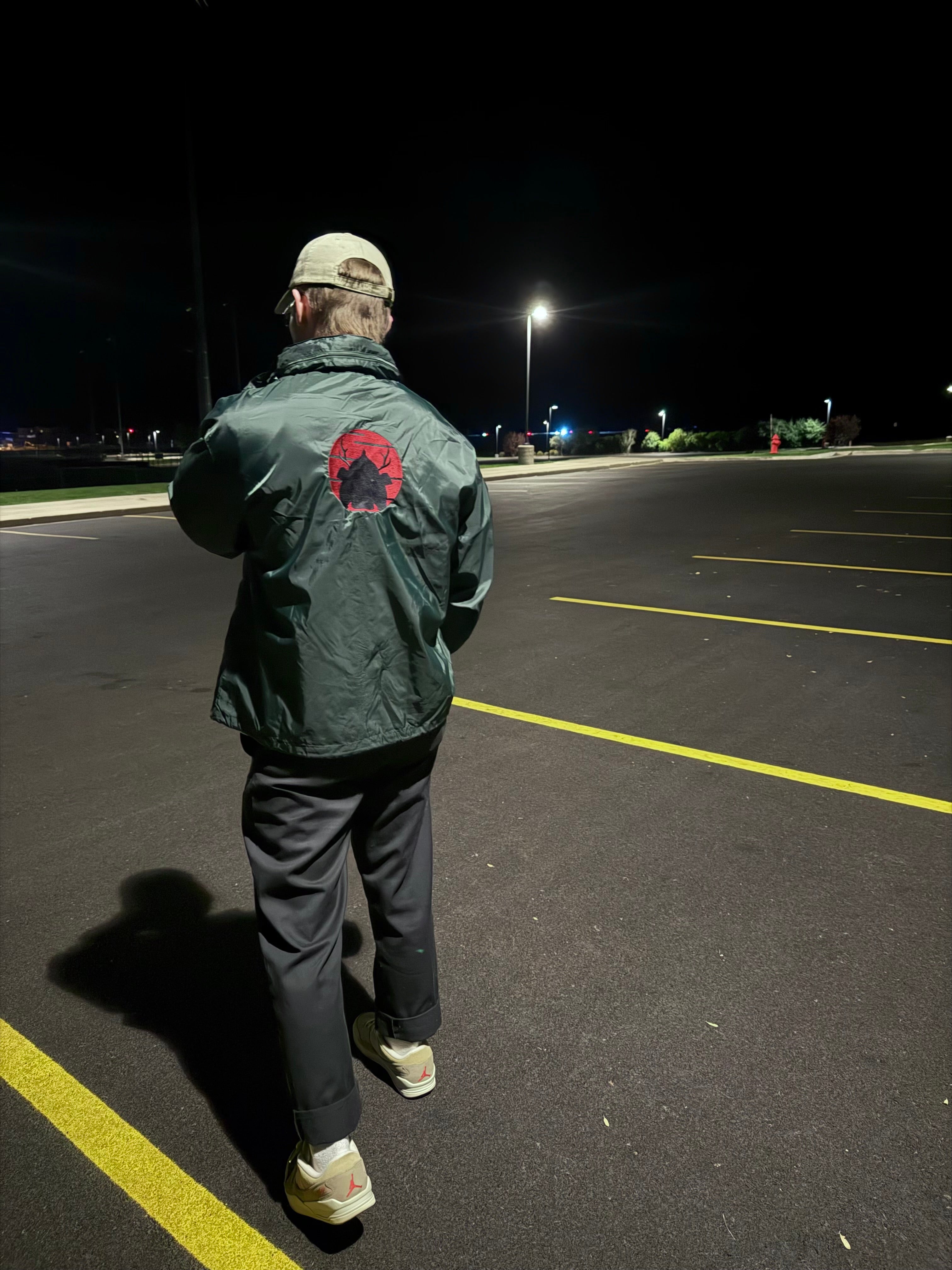Person in at night in an empty parking lot wearing a lightweight green windbreaker with Japanese red sun and samurai helmet embroidery
