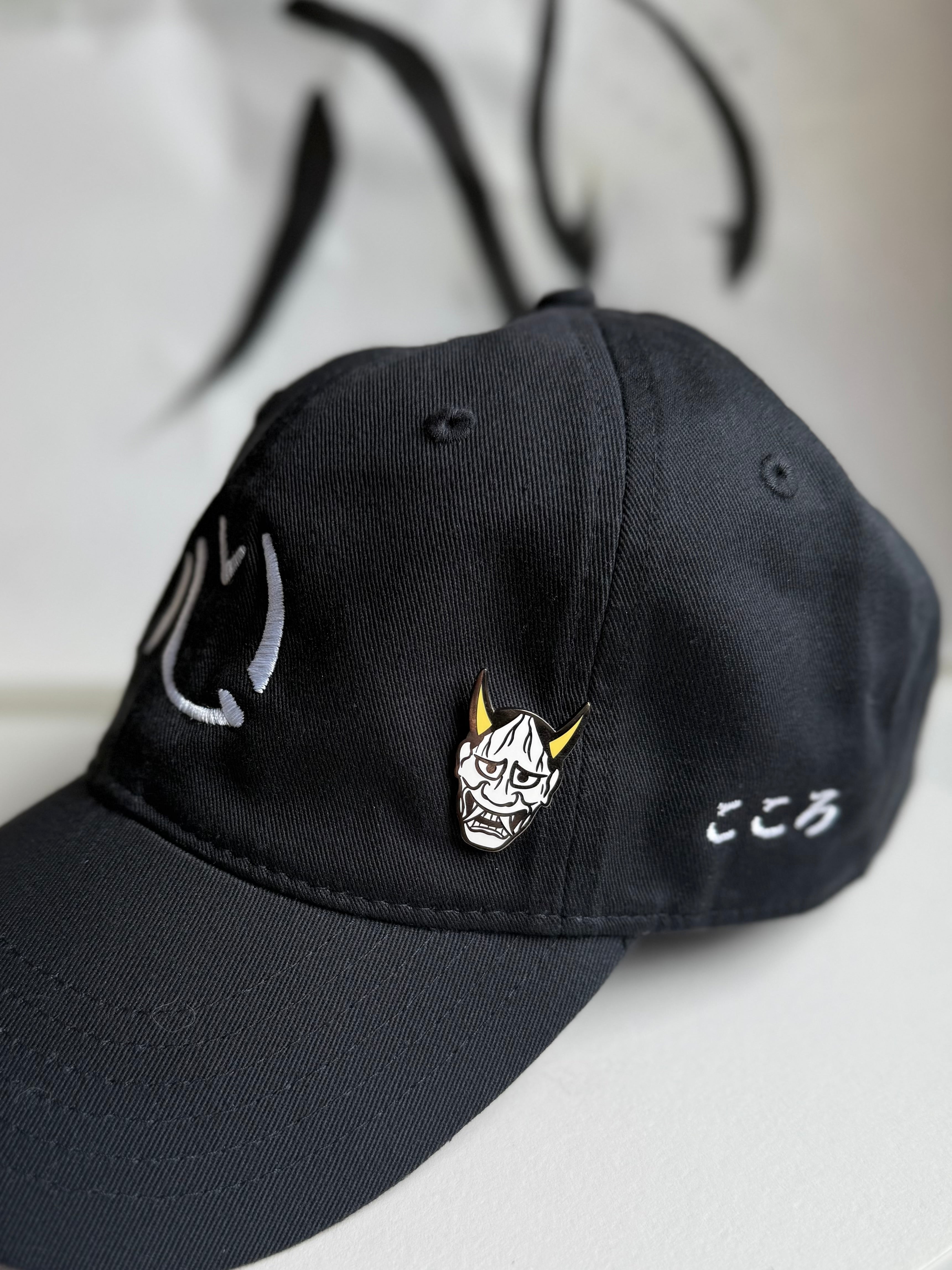 Hannya enamel pin on a black embroidered Kokoro hat with Kokoro calligraphy in the background