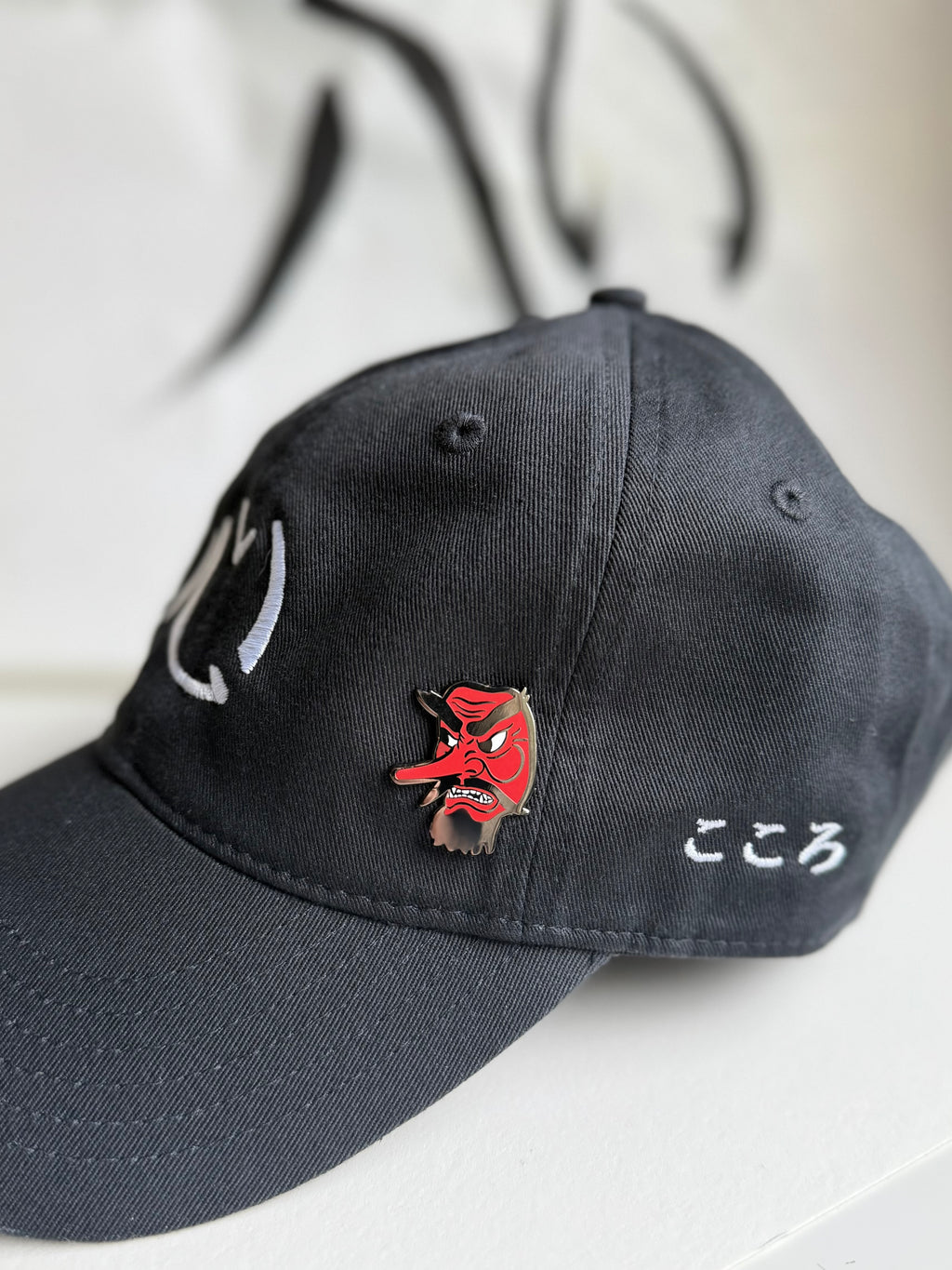Tengu enamel pin on an embroidered black Kokoro hat with Kokoro calligraphy in the background