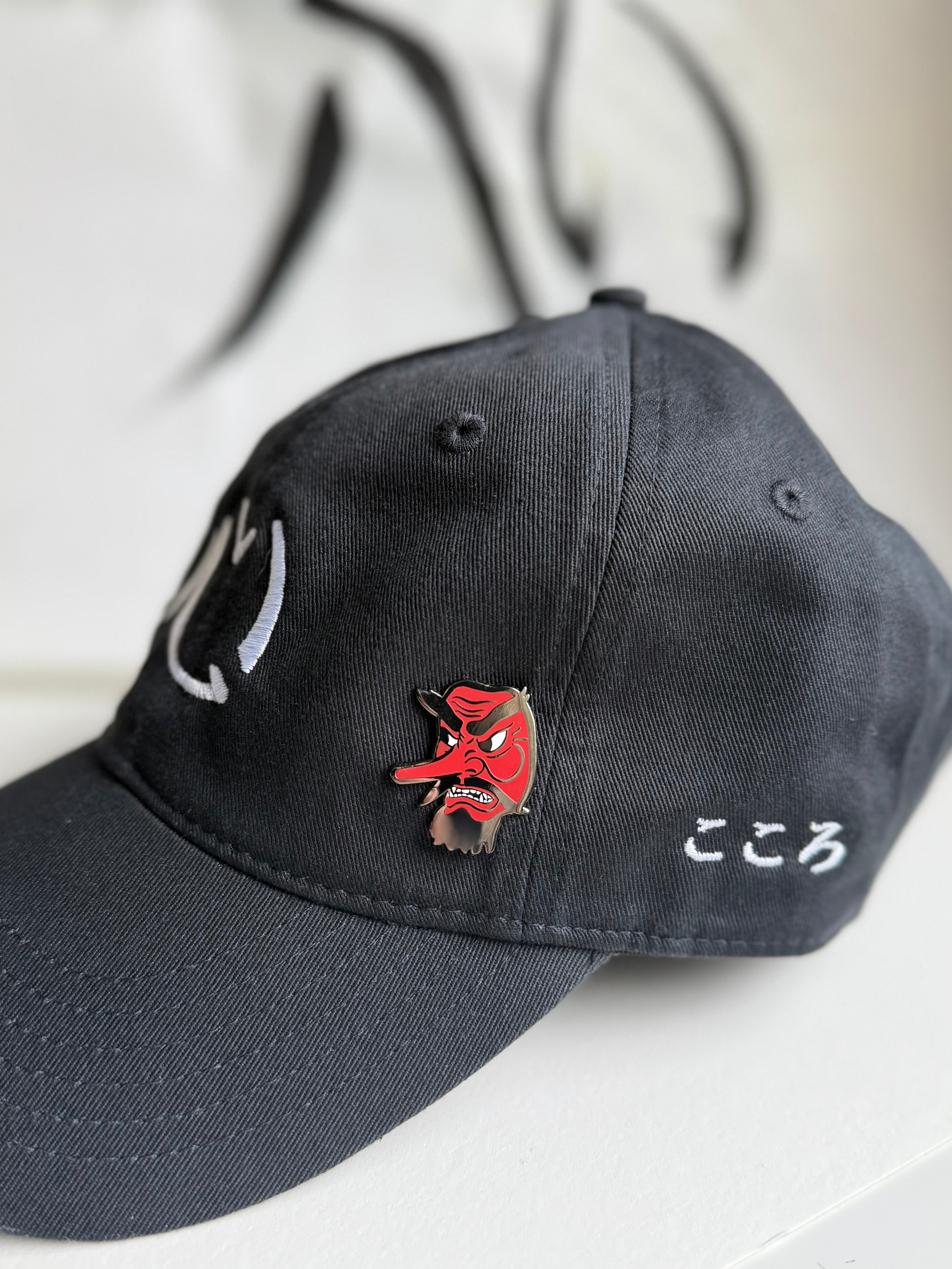 Tengu enamel pin on an embroidered black Kokoro hat with Kokoro calligraphy in the background